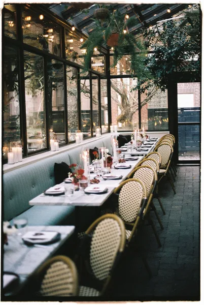Reception tablescape with long banquet table setup on a white tablecloth, taper candles and hurricane holders under string lights in a glass conservatory