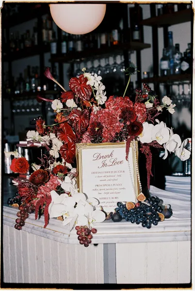 Bar wedding decor with wedding bar styling featuring a framed cocktail menu sign beside orchids, anthurium, and fruit on the counter, with liquor shelves behind