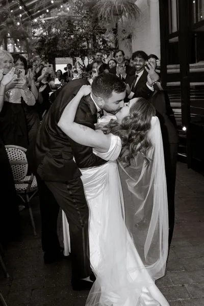 Wedding kiss portrait of groom dipping the bride as guests cheer and film on phones under string lights in a greenery-filled reception space