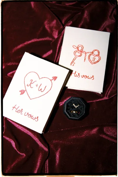 Wedding vow books with his and hers vows in handwritten red ink, heart and key illustrations, plus rings and box on burgundy satin backdrop