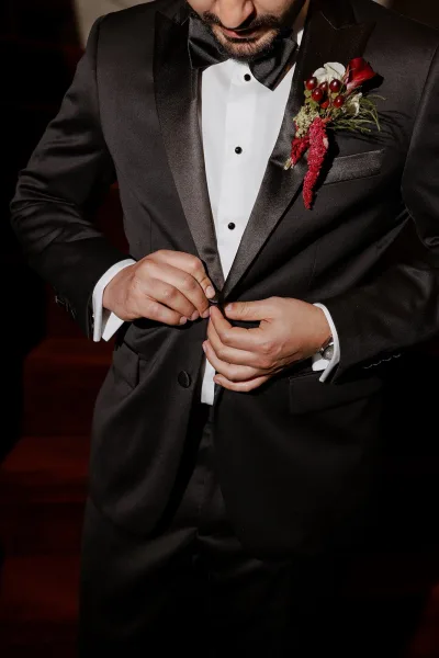 Groom getting ready, buttoning a black tuxedo with satin lapel and red boutonniere in a dark indoor setting, cufflinks visible