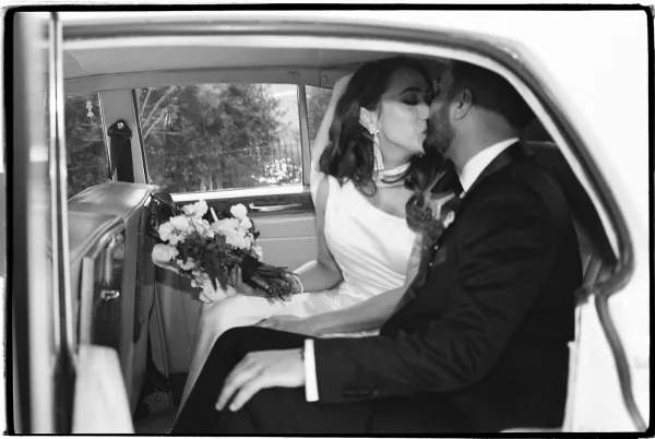 Wedding kiss portrait of bride and groom kissing in a vintage car backseat, bride holding bouquet with veil, trees through window