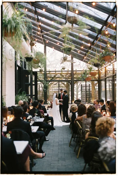Wedding ceremony with bride and groom vows under string lights in a glass greenhouse, officiant at the altar as guests watch quietly