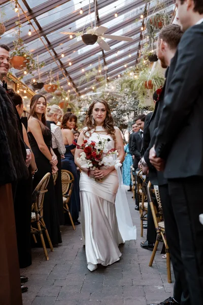 Wedding processional with bride walking down aisle holding a red and white bouquet, veil trailing in a glass-roof greenhouse with string lights
