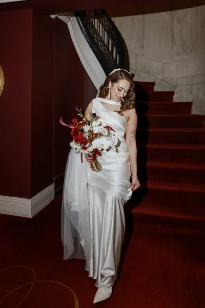 Bridal portrait of a bride on staircase in a satin gown with draped neckline, long veil, and orchid bouquet against red walls