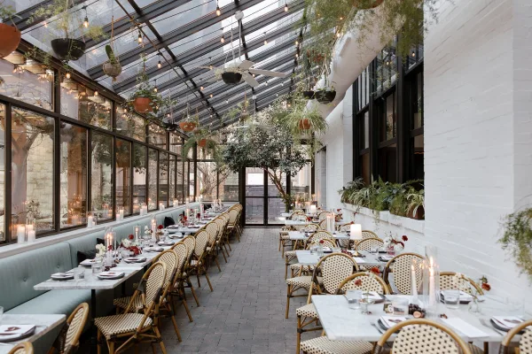 Reception tablescape with long wedding reception tables on marble, bistro chairs, white place settings, taper candles, and string lights under a glass ceiling