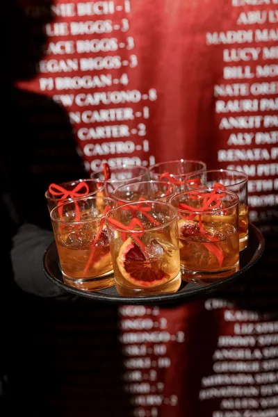 Signature cocktails on a tray in rocks glasses with ice and citrus slices, red stirrers and ribbon bows, beside a seating chart sign on dark backdrop