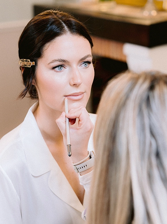 Bridal makeup as a wedding makeup artist applies lip liner, bride in a robe with a hair clip in a softly blurred indoor room