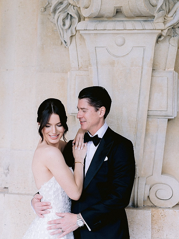 Couple portrait of bride and groom embrace, her hand on his chest showing wedding rings, against carved stone architecture backdrop