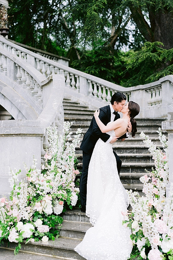 Wedding kiss portrait of bride and groom kissing in a dip on a stone staircase, her lace gown and his tux framed by rose florals and greenery