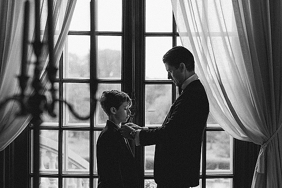 Groom getting ready adjusts his bow tie in a tuxedo by a large window, daylight streaming through panes in a quiet room
