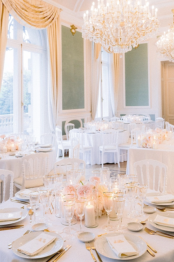Reception tablescape with round wedding tables in white linens, blush floral centerpieces and taper candles beneath a crystal chandelier in a ballroom