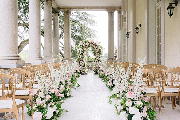 Ceremony setup with an outdoor ceremony aisle lined in blush and white flowers, leading to a floral arch on a columned veranda