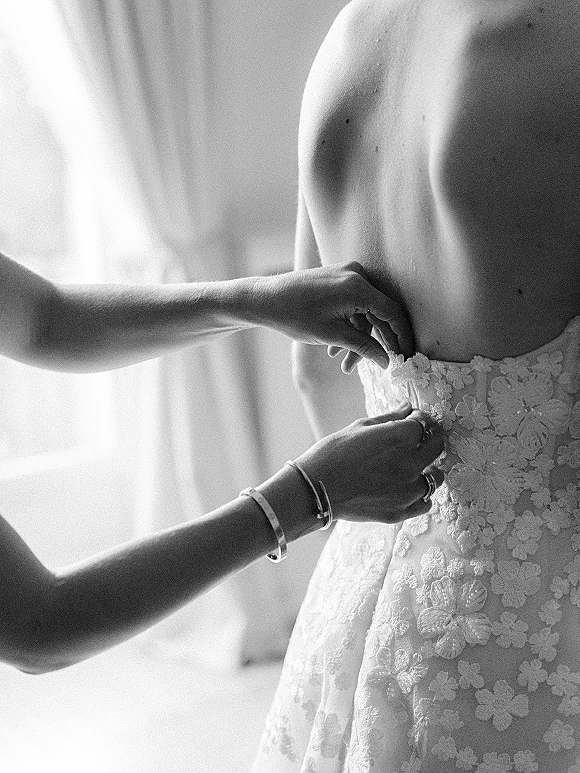 Wedding dress bustle being fastened on a strapless lace gown with floral appliqué, rings and bracelet, in window light by sheer curtains