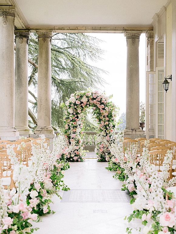 Ceremony aisle decor with a floral arch ceremony of blush and white flowers and greenery, lined with gold chairs on a stone terrace