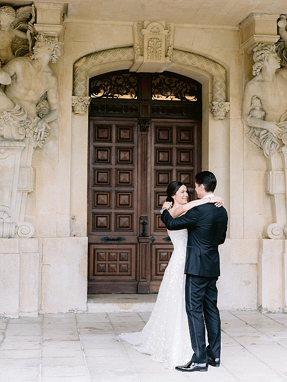 Couple portrait of bride and groom embrace beneath an ornate stone archway by wooden double doors, her strapless gown and train flowing