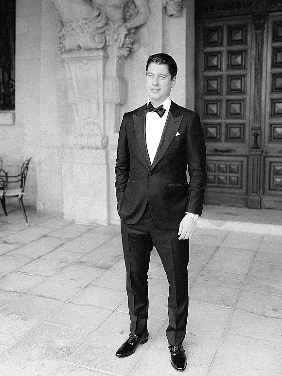 Groom portrait in a black tuxedo with bow tie and pocket square, hands in pockets in a stone courtyard by an ornate wooden door
