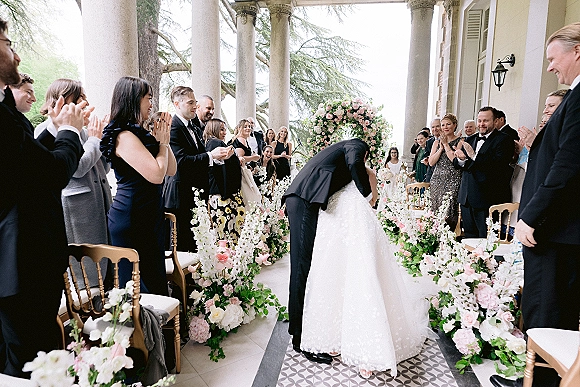 Ceremony kiss as bride in a wedding dress and groom in tuxedo embrace beneath a rose floral arch on a columned terrace aisle