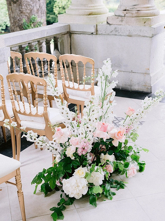 Ceremony aisle decor with an aisle floral arrangement of roses, hydrangeas and greenery beside wood chairs on a stone terrace with balustrade