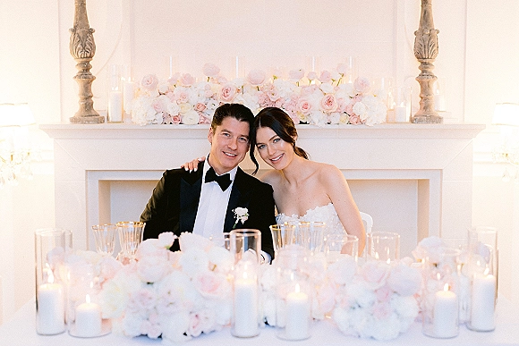 Bride and groom portrait at a candlelit sweetheart table, bride in strapless dress leaning on tuxedoed groom by a white fireplace mantel
