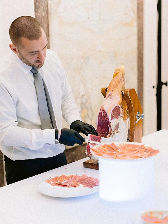 Wedding catering jamon carving station with gloved attendant slicing a cured ham leg on a wooden stand against a stone wall indoors