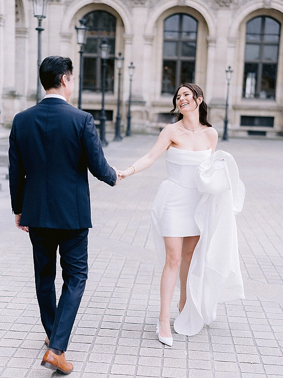 Couple portrait of bride and groom holding hands as she leads him in a stone courtyard, her strapless short wedding dress and pearl necklace catching light