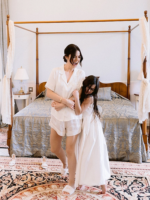 Bridal getting ready in a satin robe and slip dress, wearing white slippers beside a canopy bed in a bright bedroom suite