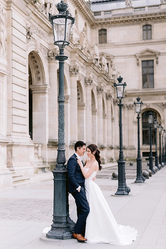 Couple portrait of bride and groom pose by a stone building facade, her strapless gown and necklace beside his navy suit under street lamps