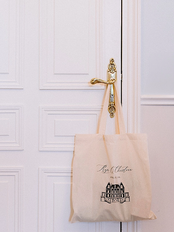 Wedding welcome bag canvas tote with printed monogram and illustrated venue graphic hanging on a gold door handle against a white paneled door