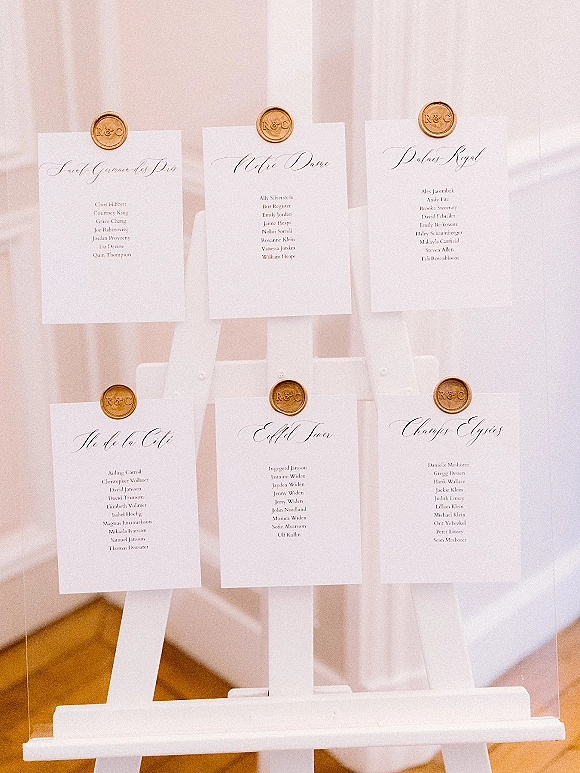 Wedding seating chart with white calligraphy cards and gold wax seals on an acrylic panel easel, set against white drapery on wood floor