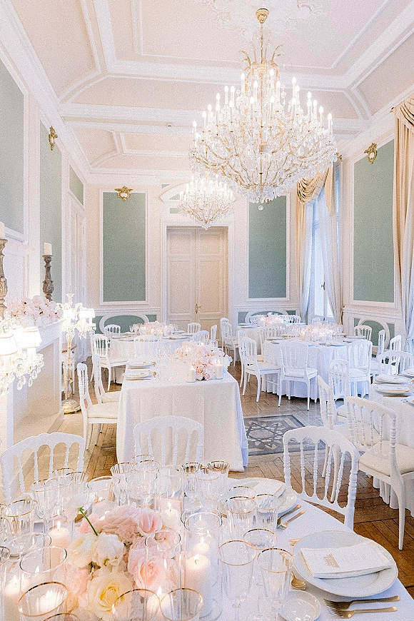 Reception tablescape with wedding reception tables set in white linens, blush floral centerpieces, candles, and crystal stemware under chandeliers