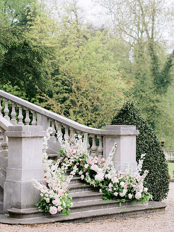 Wedding staircase florals with ceremony entrance florals in pink and white roses, hydrangeas, and greenery along stone steps and balustrade in a garden estate