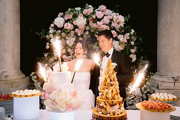 Wedding cake cutting by bride and groom as sparklers flare beside a champagne tower, framed by roses and greenery near stone columns