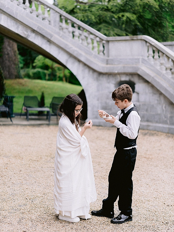Wedding kids moment as a flower girl and ring bearer sit on a stone staircase eating dessert, her eyeglasses catching light in a garden courtyard