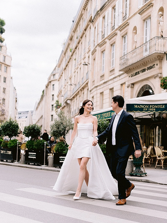 Couple portrait of bride and groom holding hands, crossing a city street in a short dress with train, heels, and navy suit near a cafe storefront