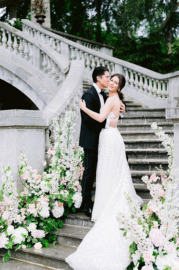 Couple portrait of groom kissing bride’s cheek on a stone staircase, her wedding dress train draped beside lush floral arrangements