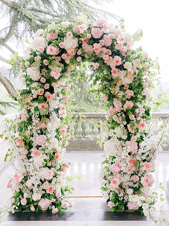 Wedding ceremony arch with pink and white flowers, one blush rose accent, and trailing greenery on an outdoor terrace with hills beyond