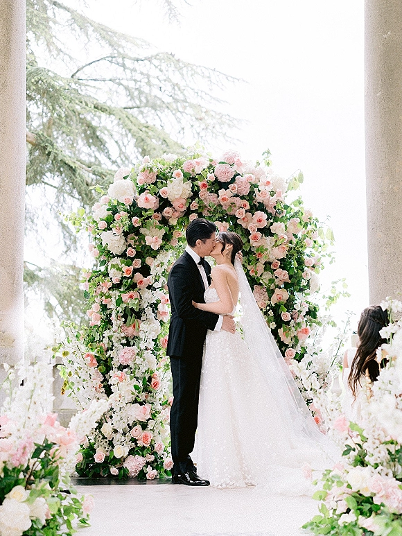 Wedding kiss portrait of bride and groom kissing under a round floral arch with roses and greenery, framed by stone columns and trees outdoors
