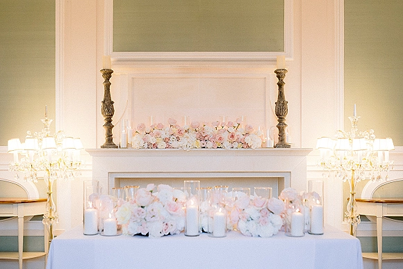 Wedding sweetheart table with rose arrangements and pillar candles in glass hurricane vases, set before a fireplace mantel garland backdrop