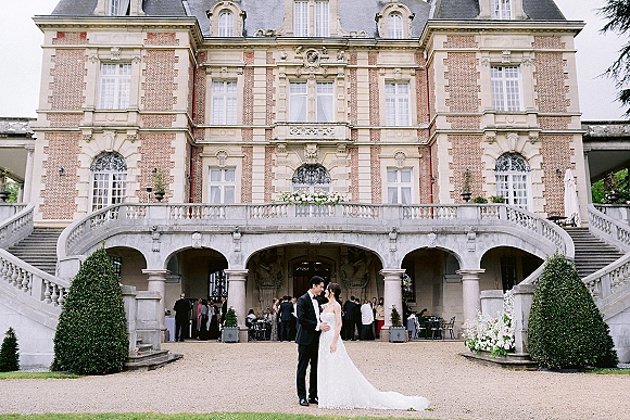 Couple portrait of bride and groom embracing in formal attire before a historic mansion staircase, with floral arrangement and guests behind