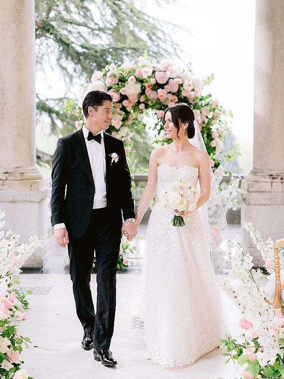 Couple portrait of bride and groom holding hands, her veil and bouquet flowing as they walk beneath a floral arch on a stone terrace