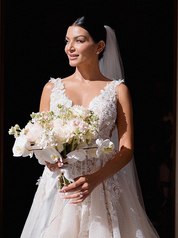 Bridal portrait of a bride holding bouquet with white orchids and blush flowers, wearing a veil and lace dress in a dark doorway