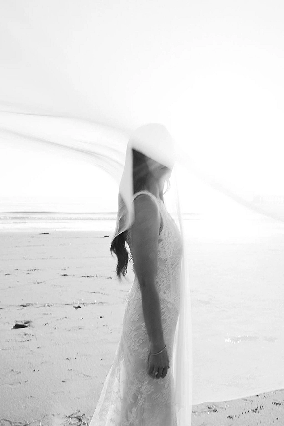 Bridal portrait of a bride in veil, lace wedding dress and bracelet, looking down as her long veil blows by the ocean shoreline