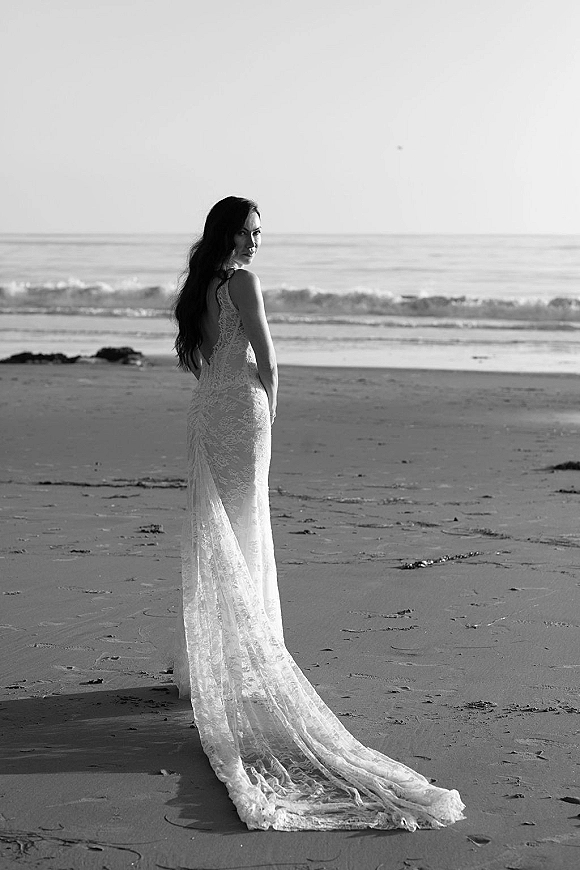 Bridal portrait on a sandy beach, bride looking over shoulder in a low-back lace wedding dress with long train, ocean waves behind