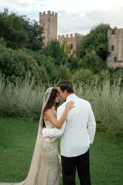 Couple portrait of a bride and groom embracing, her long veil trailing as they stand by castle walls and garden shrubs under a cloudy sky.