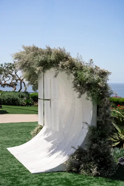 Wedding seating chart and escort card display on a sculptural backdrop, set on a lawn with ocean views beneath a clear blue sky.