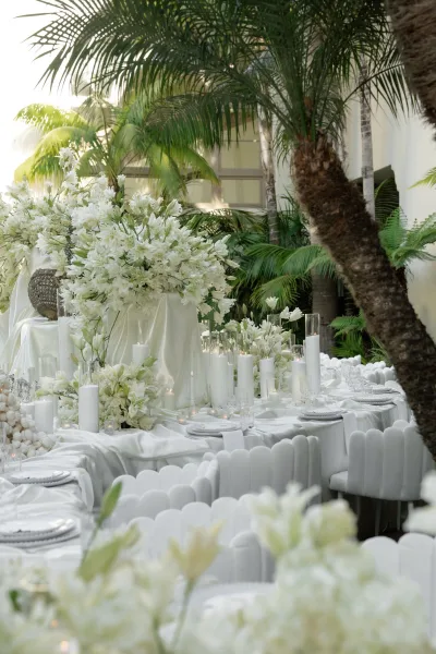 Reception tablescape in all white reception decor, accented by lilies, in a palm-lined courtyard with tropical greenery.