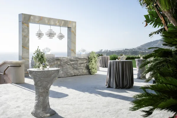 Cocktail hour setup for an outdoor cocktail hour featuring a marble arch, with an ocean view backed by palm trees and a hillside.