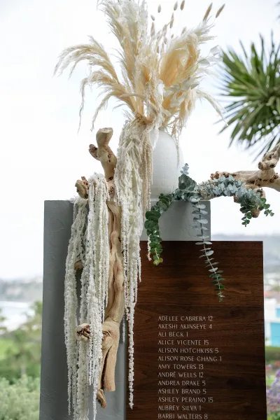 Wedding seating chart and table assignments sign trimmed with pampas grass, displayed outdoors against open sky, a palm tree, and distant greenery.