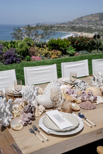 Reception tablescape styled for beach wedding table decor, featuring seashells along the runner, set by an ocean coastline with garden lawn.